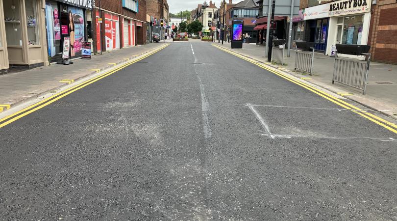 A smooth and freshly resurfaced road with bold double yellow painted lines either side and businesses lining the road on both sides.