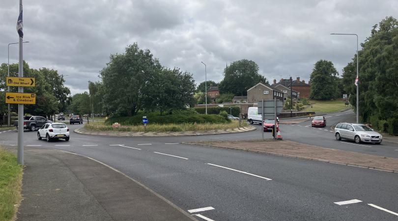 A roundabout with a smooth newly laid road with freshly painted markings and cars driving on the roundabout.