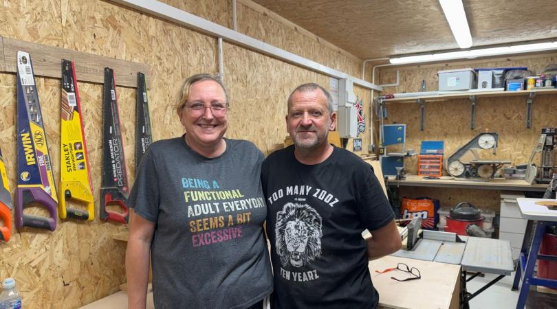 A woman and a man inside a workshop surrounded by tools, including hand saws hanging up on the wall behind them.