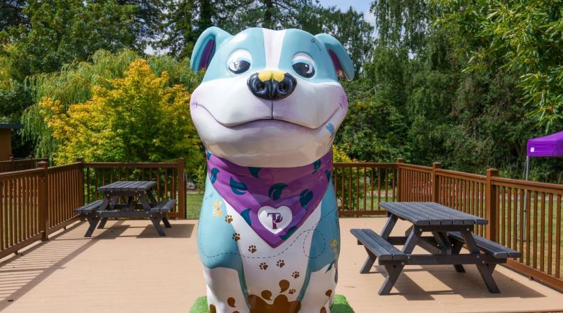 A large turquoise sculpture of a Staffordshire Bull Terrier with colourful artwork painted onto it, including a purple bandana around its neck. It is placed in an outdoor courtyard with trees and picnic benches behind.