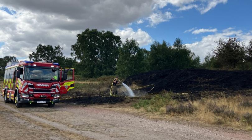 A red fire engine pulled up at the side of the road. A firefighter is holding onto a hosepipe, which is connected to the fire engine and hosing down a blackened area of heathland in the countryside. Above, the sky is bright blue with some grey clouds gathering to the left.