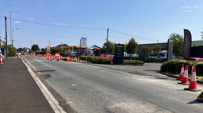 An empty road that has had some patch repairs along the sides, with some orange traffic cones closing off the right lane. To the right of that are some shops.