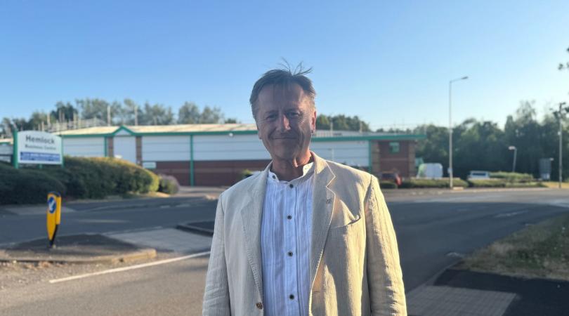 A man standing front and centre, smiling at the camera, whilst stood next to a road.