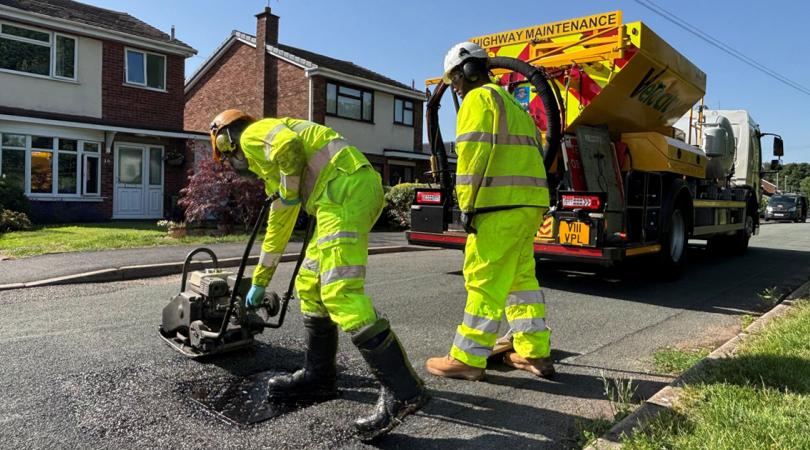 Two highways workers wearing bright yellow jackets and trousers. They're using a wacker machine to flatten down a pothole they have just filled. A big yellowy-orange highways truck can be seen in the background. It is a very hot and sunny day.