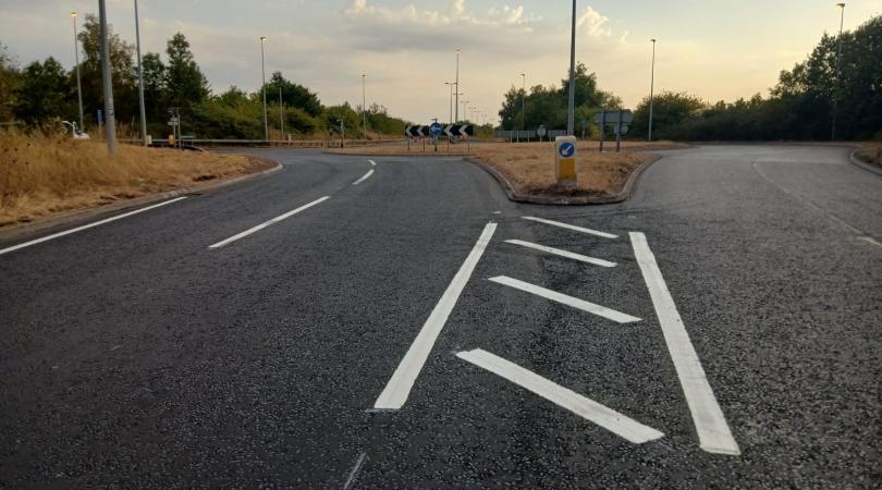 Image shows a lovely, flat, newly resurfaced  road leading up to a roundabout. There are hatch markings and white lines visible. The image is taken late evening so the sun is low and basking everything in an ethereal glow.