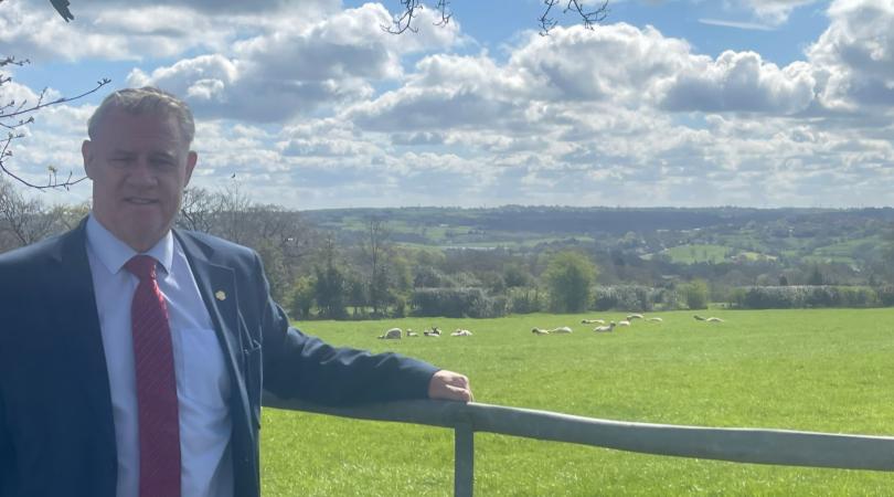 Anthony screen in the countryside, leaning on a fence with a field and sheep behind him