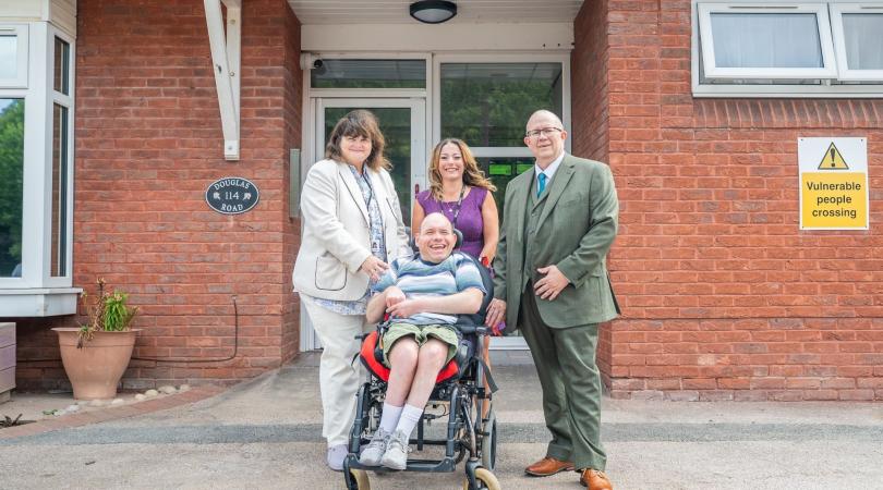 Two smartly dressed women and a man standing behind a man in a wheelchair. They are all smiling as they are stood outside the entrance to a brick building.