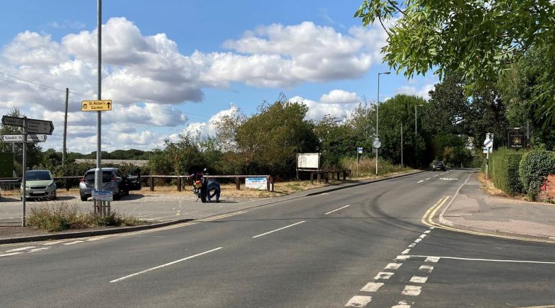 A four-way junction in Burton, with a bus stop and parking area to the right of the road. It is a sunny day.