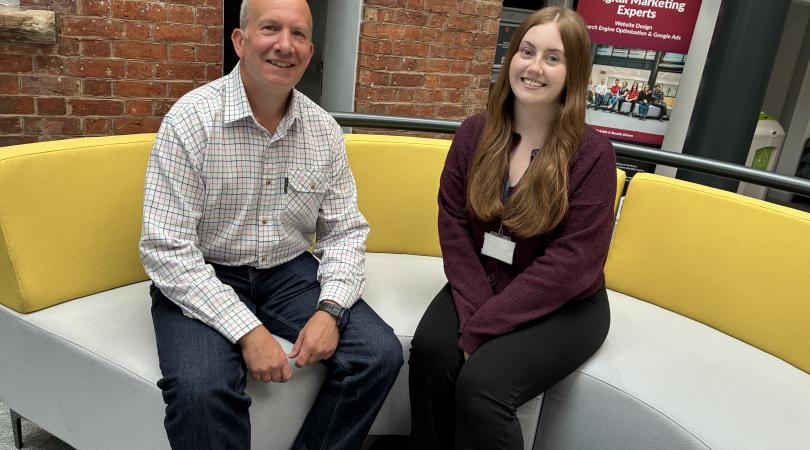 A smartly dressed smiling man and woman sitting down on a modern curved yellow and grey sofa.