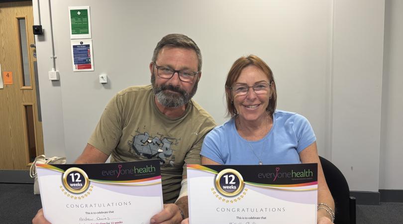 Andrew and Michelle sitting at a table holding certificates from Everyone Health recognising them for being smoke-free for 12 weeks. The certificates are displayed towards the camera in an indoor meeting room setting.