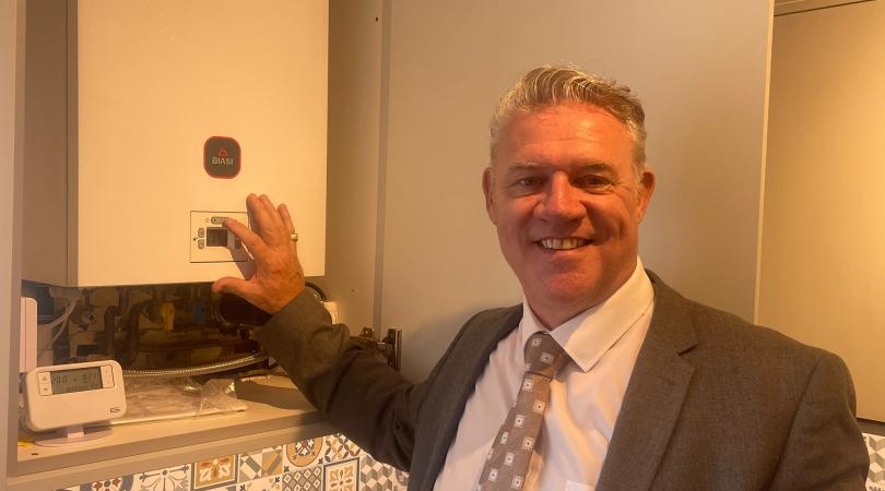 Anthony Screen stands beside a domestic boiler, pointing to the controls.