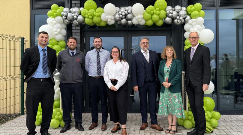 Group of seven people standing outside Greywood school entrance beneath a green, white and silver balloon arch during a school opening or celebration event.