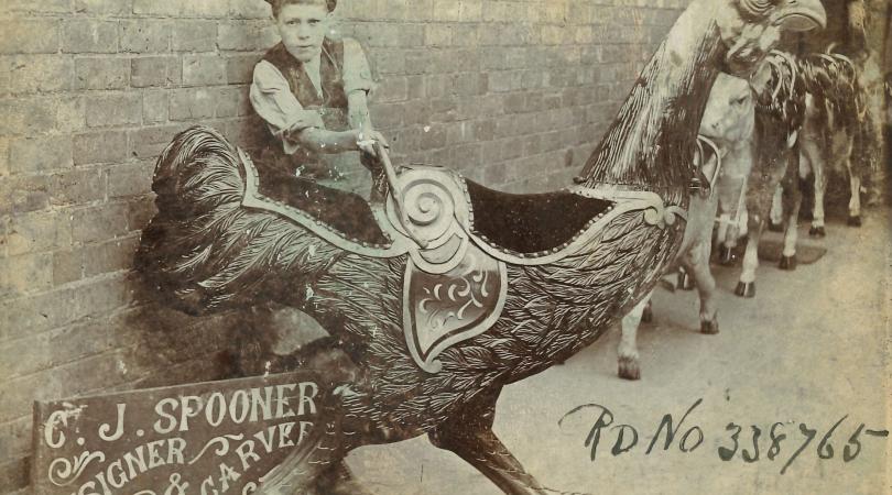 Historical photograph showing a young boy posing beside a carved fairground cockerel manufactured by C.J. Spooner.
