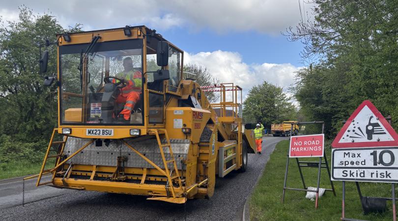 Road resurfacing works on a rural road, with a yellow paving machine, road workers in high-visibility clothing, and warning signs advising of no road markings and a 10mph maximum speed.