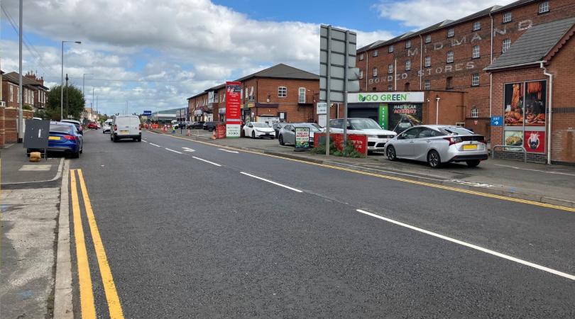 A smooth newly resurfaced road with freshly painted double yellow lines running along the side of it and several vehicles driving along the route. It is a bright day with blue sky peeking through the clouds in the sky.