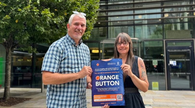 A smiling man in a checkered t-shirt standing with a woman with dark hair and a dark dress in front of a building outside and holding a poster promoting the 'Orange Button' scheme.