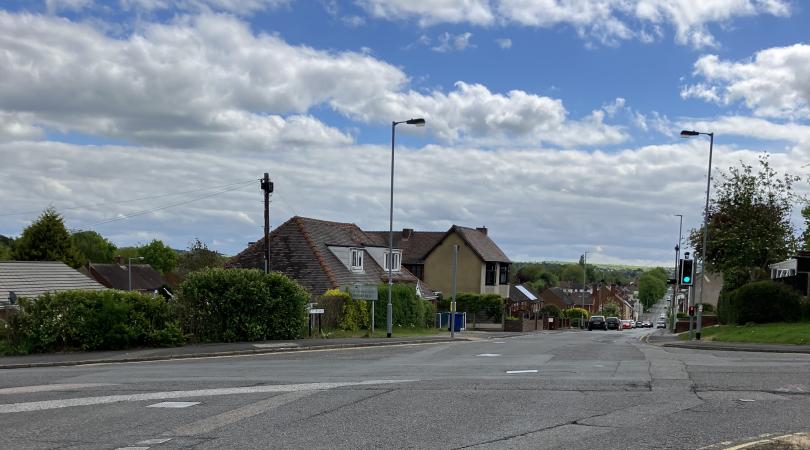 A picture of a crossroads with houses in the middle distance on a cloudy day. The road looks tired and in need of refurbishment.