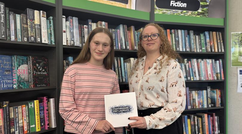 Lola (left) and Cherry (right) stand in front of bookshelves in Stafford Library, holding a Staffordshire Poet Laureate folder beneath the library's fiction section signage.