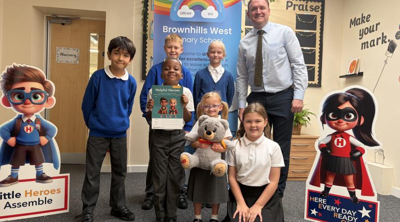Pupils and staff at Brownhills West Primary School pose with a Little Heroes resource booklet and teddy bear, standing between two superhero character displays in a school classroom.