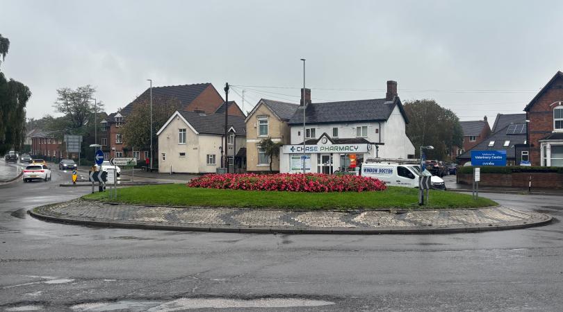 A picture of a roundabout on a grey, rainy day. The roundabout has red flowers on it. There is a white house in the background.