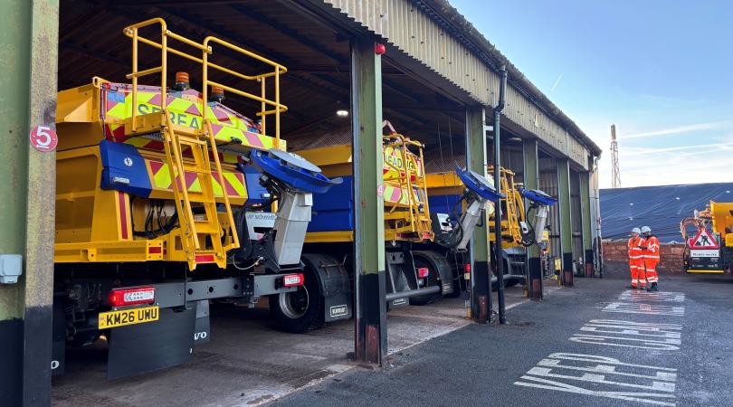 A row of three gritting machines in a depot shed. You can see the rear of the gritters, and the spinner where the salt comes out. There are two men in bright orange in the far right of the photo.