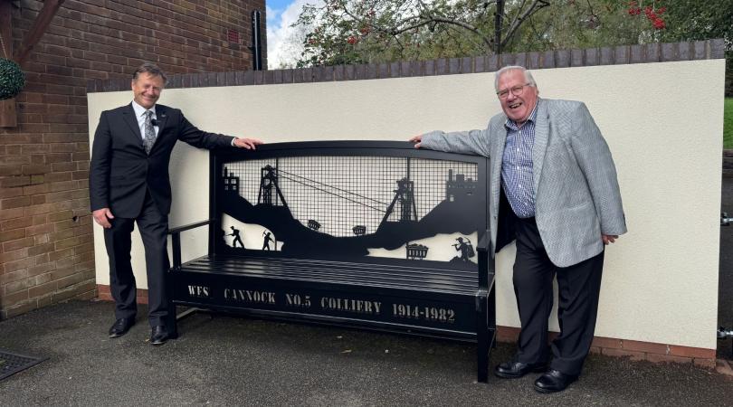A black steel bench with mining designs on the backrest. There are two men, smartly dressed in suits, smiling and leaning on each side of it.