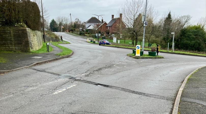 A surface-worn road approaching a roundabout. The day is overcast.