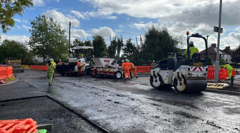 A group of highways workers, wearing bright orange and yellow hi-vis and white hard hats, working on repairing a road with various bits of machinery, including a roller. The work site is surrounded by bright orange plastic fencing and there is a bright blue sky above them with some clouds.