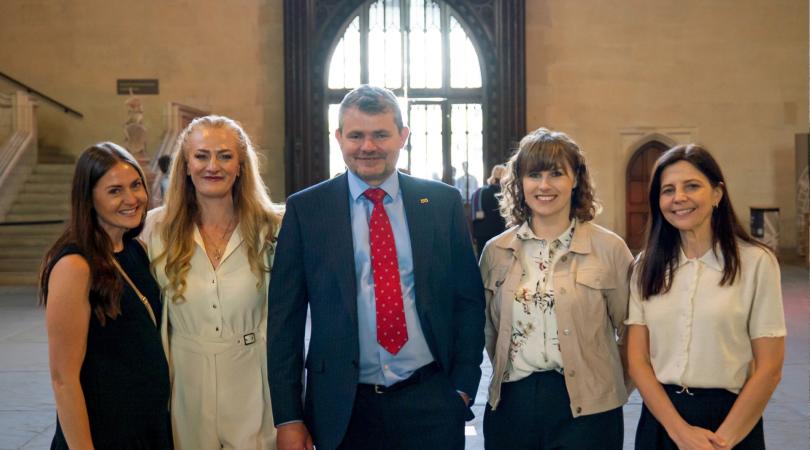 Four smartly dressed women and a man standing together and smiling at the camera. They are indoors and, behind them, is a staircase and an arched window that stretches from the ceiling to the floor.