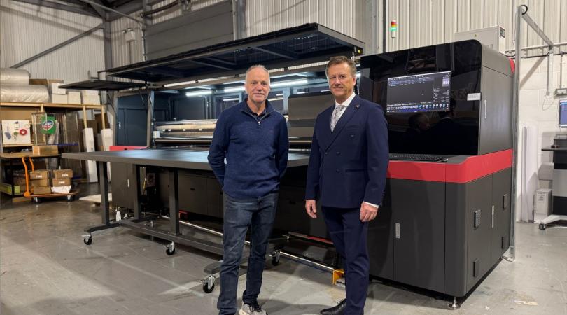 Two smartly dressed men standing in front of a large industrial printer.