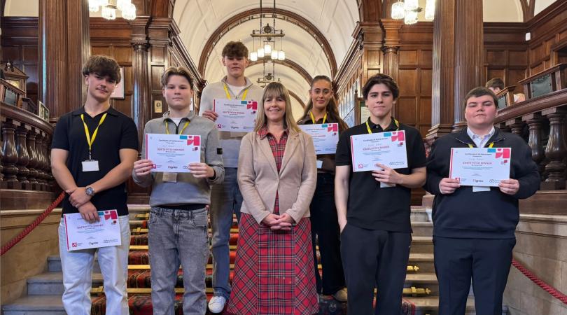 Five students facing forward, smiling, and holding certificates. A smiling woman in a red tartan dress and brown jacket is standing in the centre of them. They are indoors, standing on a wide red carpeted staircase. 