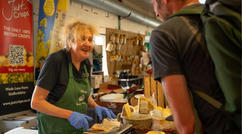 A smiling woman, wearing a green apron and blue gloves serving cheese to a male customer.