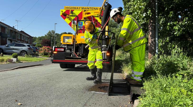 Two highways workers in bright yellow, next to a large highways machine, repairing a road.